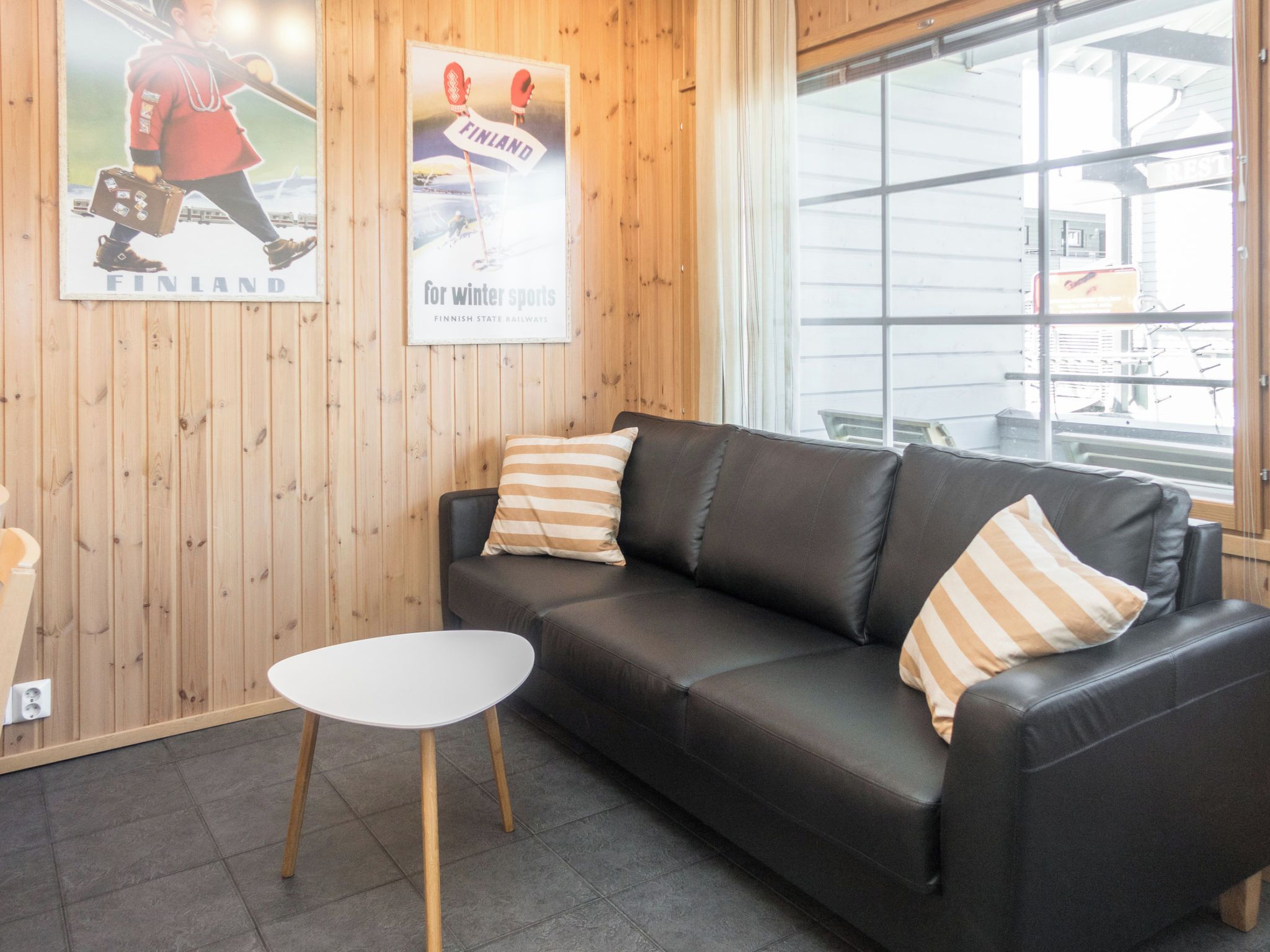 A room with a black leather sofa, two striped pillows, a small white table, wooden panel walls, and two posters advertising winter sports and Finnish railways.