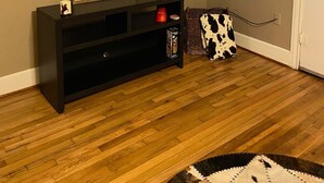 A living room with a flat-screen TV mounted on the wall displaying a streaming service interface. A dark wooden entertainment center with a 'Blessed' sign is below the TV. A cow-patterned throw rug is on the floor, and a cross is on the wall. - Cozy Downtown Home (Bryan)