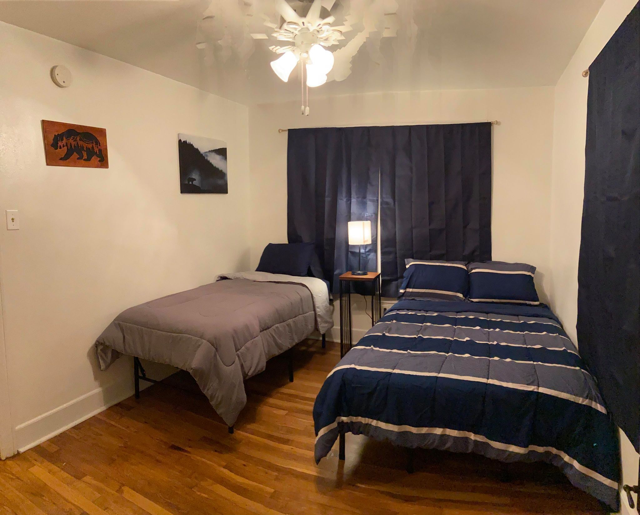 Een slaapkamer met twee bedden, een plafondventilator, een nachtkastje met lamp en twee aan de muur hangende foto's.