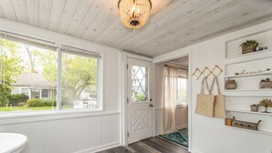 Una habitación con techo de madera, un candelabro, una puerta blanca y vistas a una casa a través de amplios ventanales.