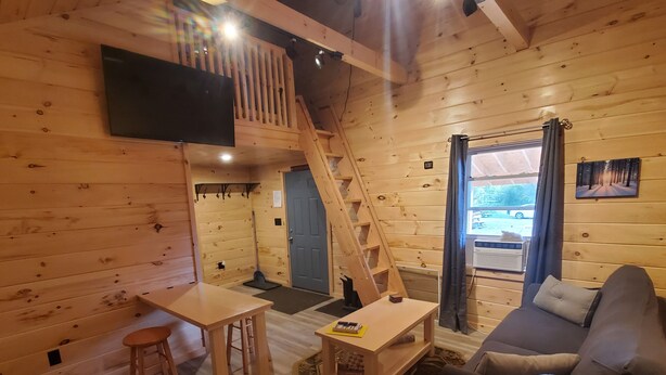 A cozy cabin interior with a wooden floor, a sofa, a table, a TV mounted on the wall, a staircase, and a window with curtains.