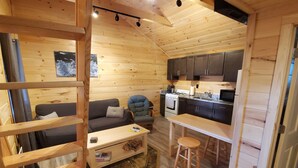 A cozy cabin interior with a wooden floor, a sofa, a dining table, a kitchen area with cabinets and a stove, and a ladder leading to a loft space.
