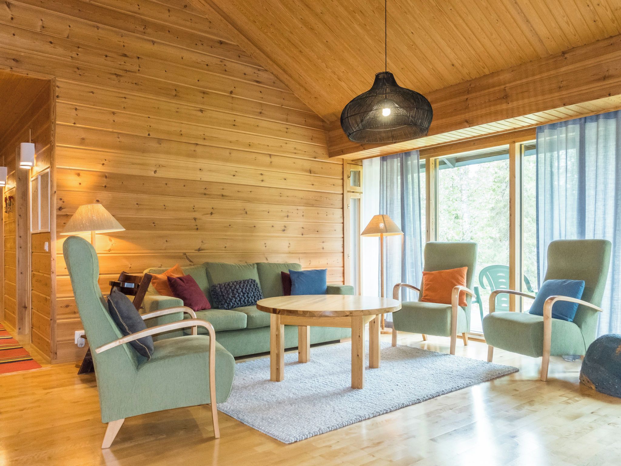 A cozy room with wooden walls and floors, a green sofa, two armchairs, a round wooden table, and a hanging light fixture.