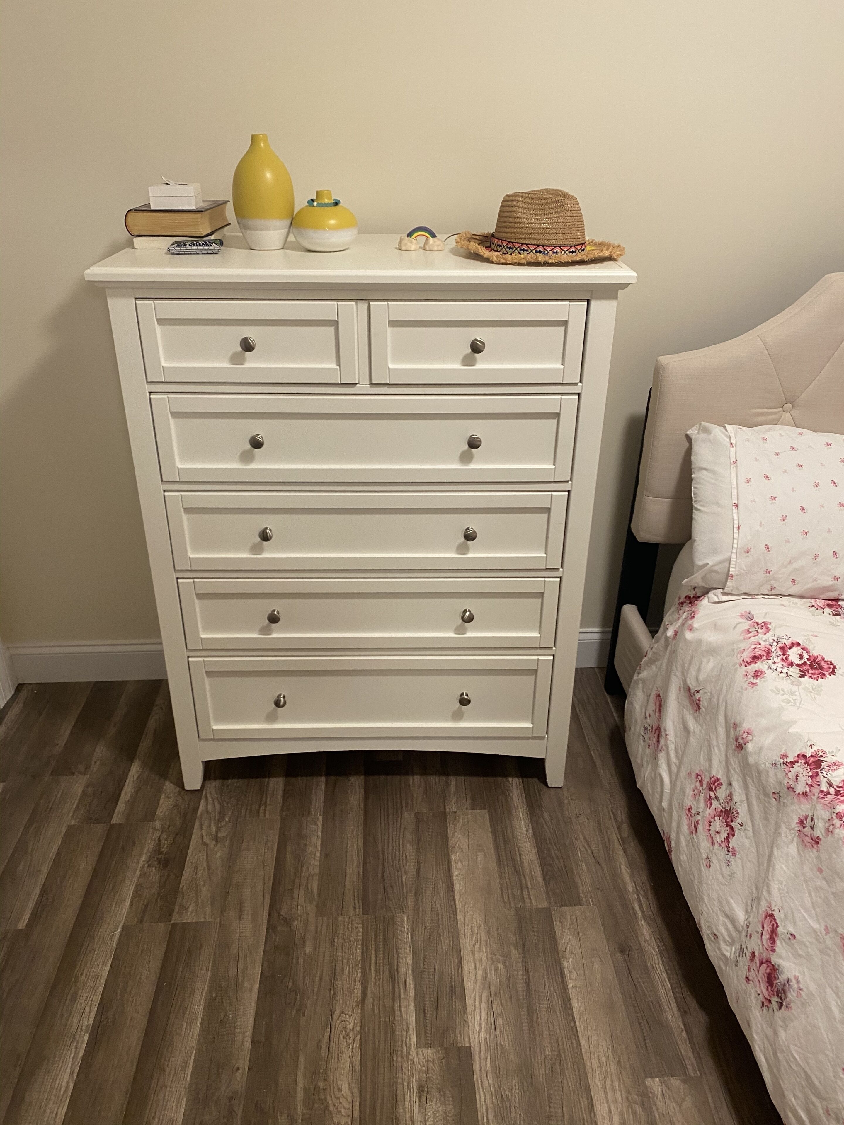 Une commode blanche avec de nombreux tiroirs, un vase jaune, un petit récipient et un chapeau de paille posés sur le dessus.