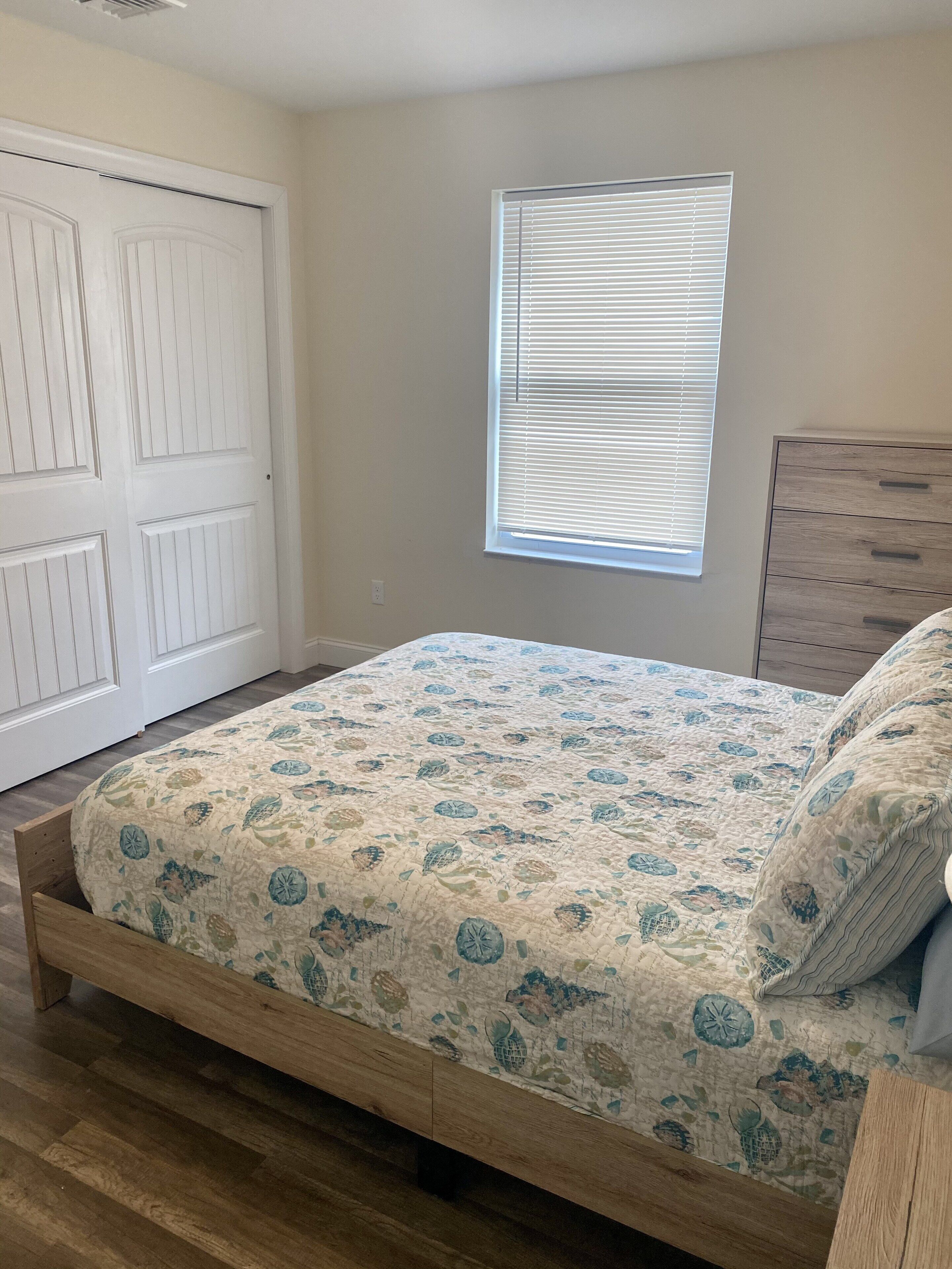 Un dormitorio con una cama de madera, una ventana con persianas, una puerta blanca y un aparador de madera.