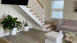 Un salon avec un escalier, un téléviseur à écran plat fixé au mur, un canapé beige, un pouf blanc et deux plantes en pot.