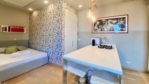 Een slaapkamer met een bed, een muur met behang met een patroon, een klein tafeltje met een waterkoker en een lijst met een foto aan de muur.
