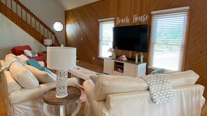 A cozy living room with a staircase, a flat-screen TV, and a sign reading 'Beach House'. - Island Haven - Hot Tub & Beautiful Water Views (Nags Head)