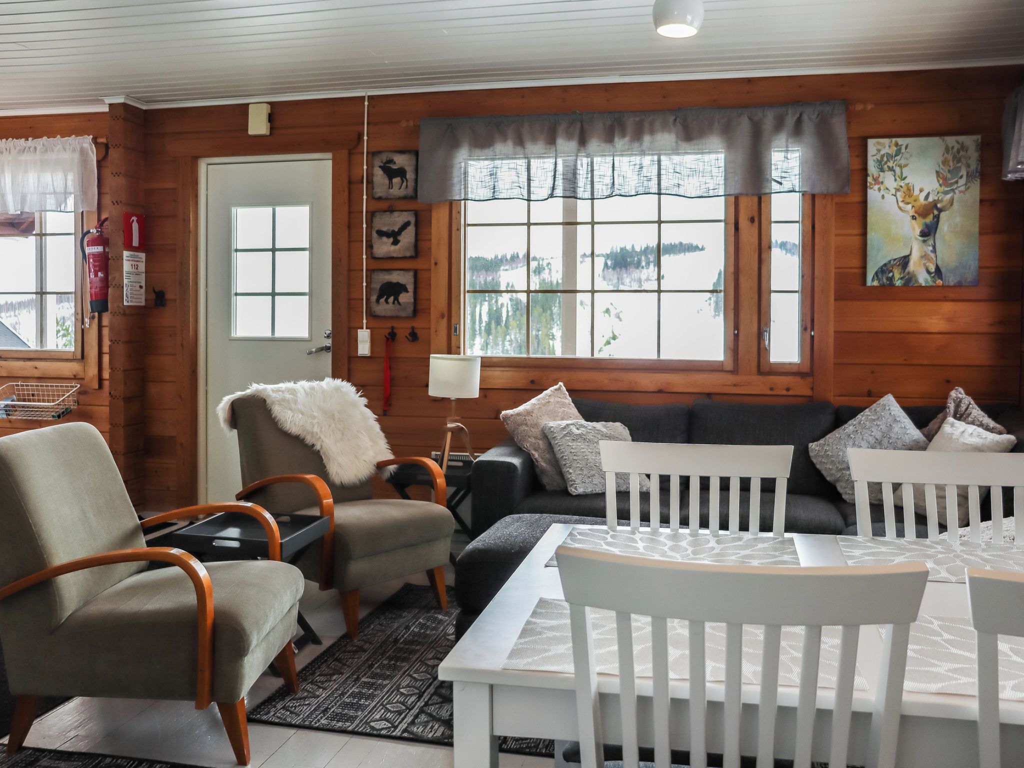 Interior acogedor de una cabaña con detalles de madera, un conjunto de sofás, una mesa de comedor y vistas a un paisaje nevado a través de grandes ventanales.