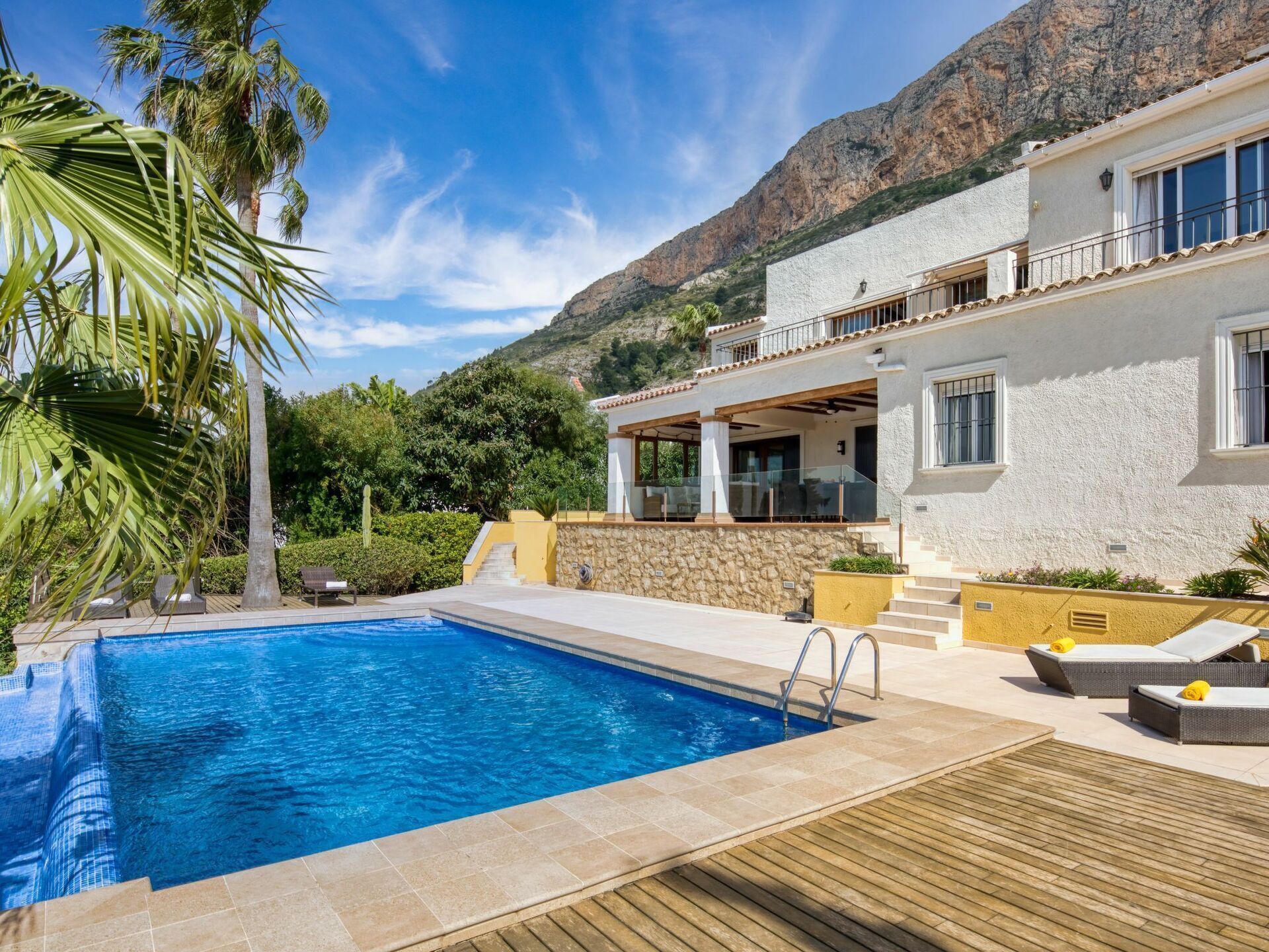Uma área à beira da piscina com uma piscina azul cristalina, um deck de madeira e um edifício branco com uma varanda com vista para a piscina.