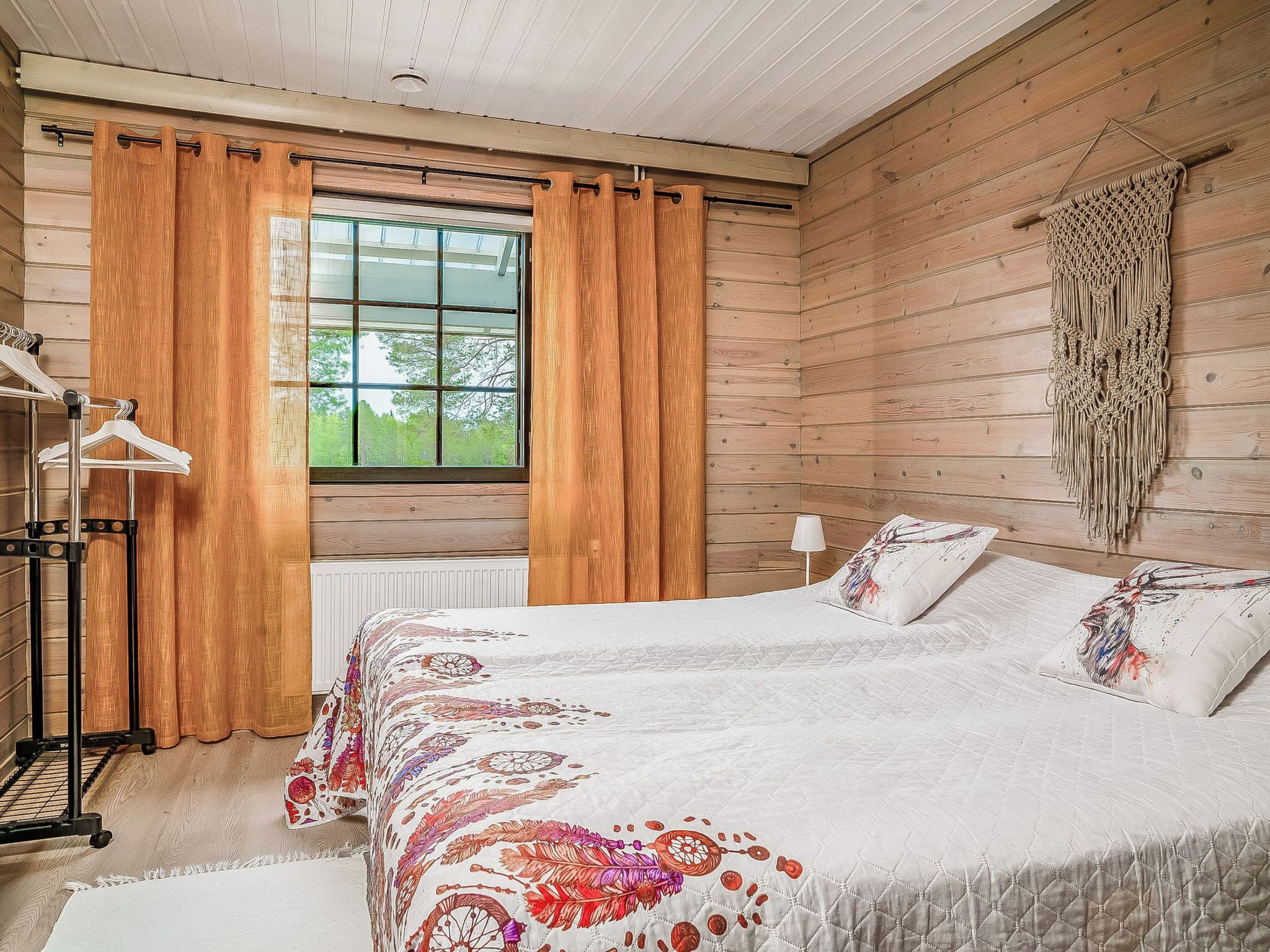 Un dormitorio con cama, paredes de madera, ventana con cortinas, radiador, un adorno en la pared y una mesita de noche.