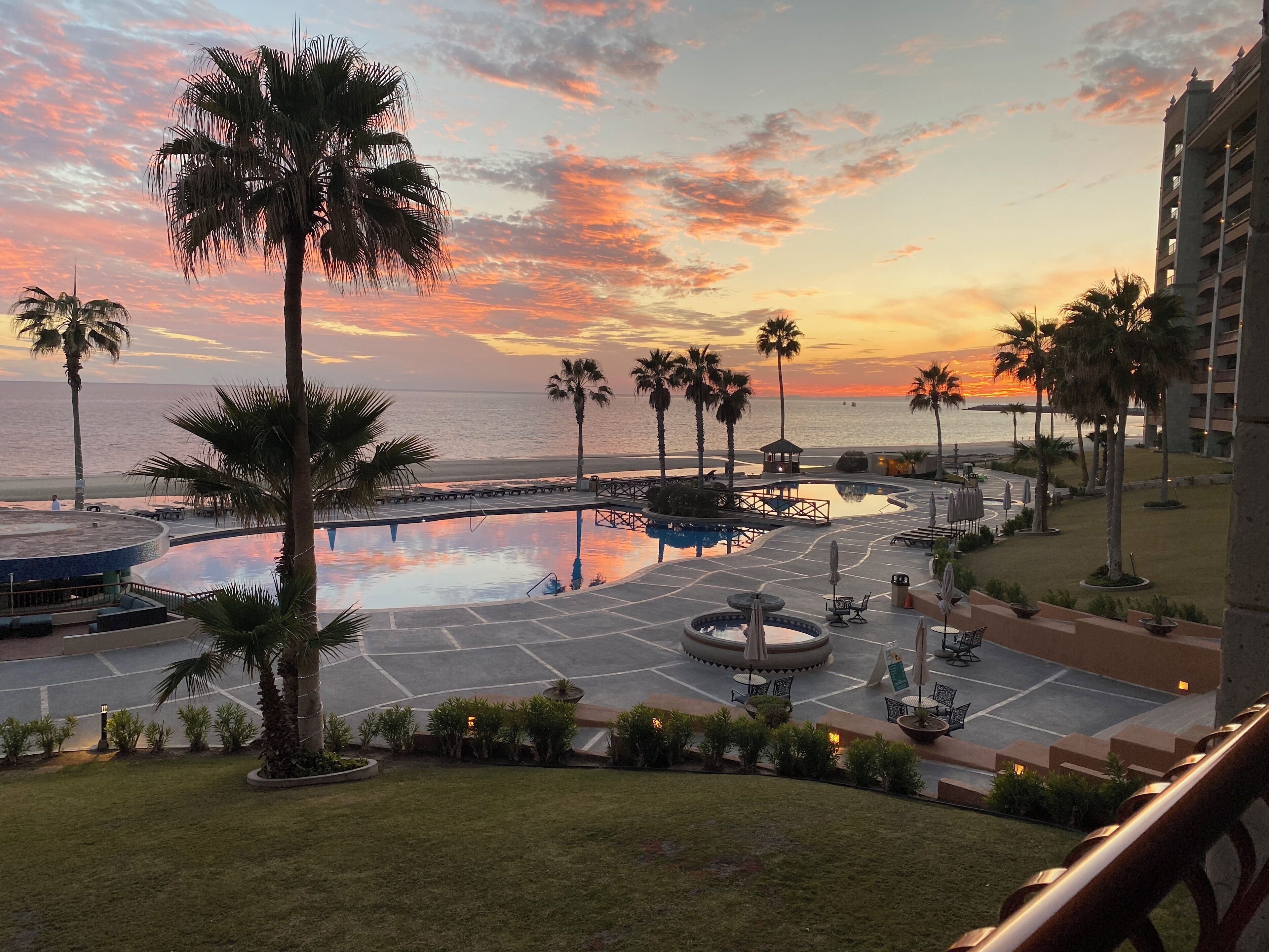 A resort pool area with palm trees, a circular water feature, and a view of the ocean at sunset.