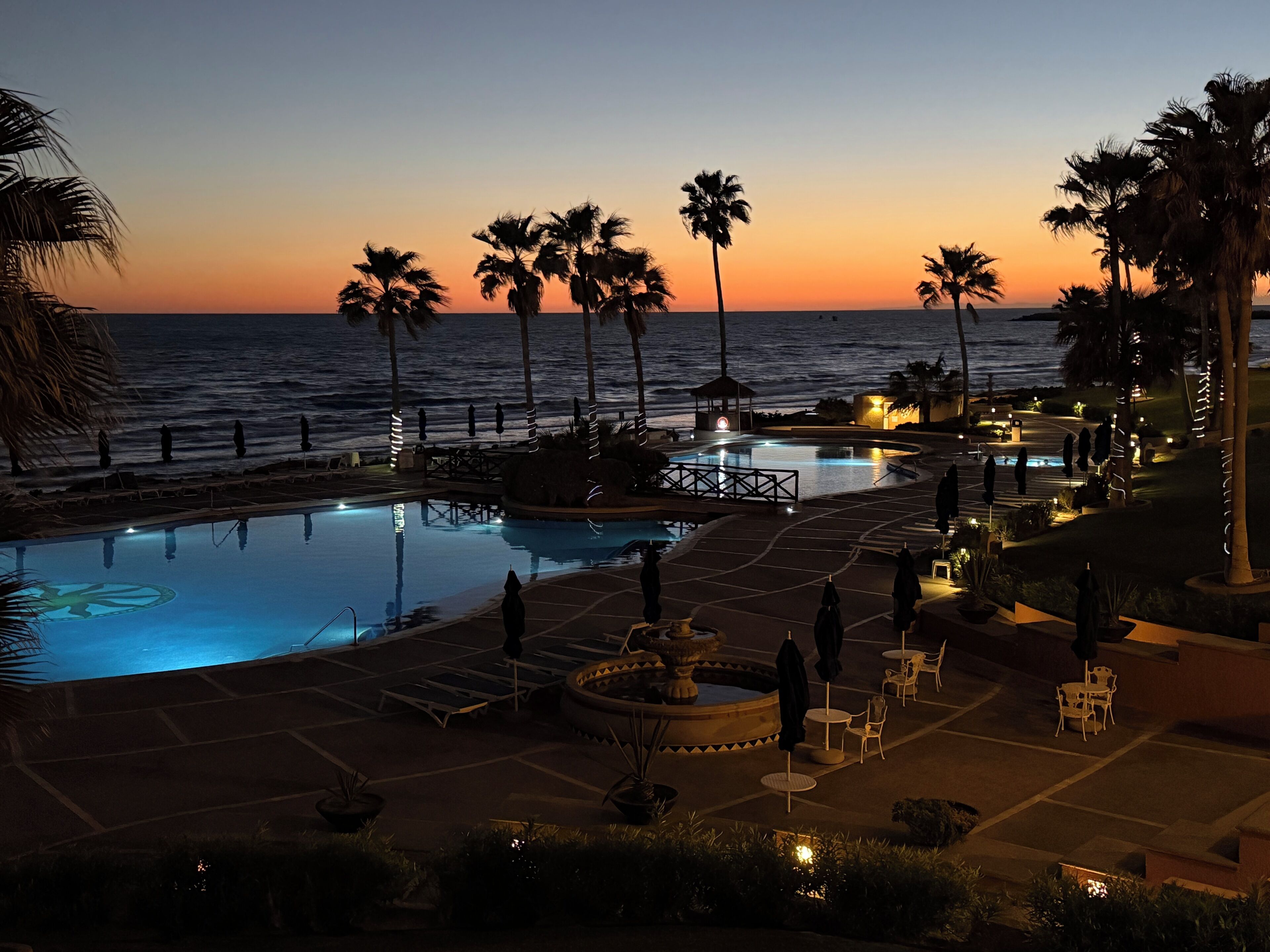 Une piscine d’un complexe hôtelier offrant une vue sur l’océan au coucher du soleil, entourée de palmiers et de chaises longues.