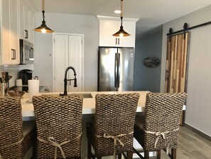 A modern kitchen with a central island, bar stools, a stainless steel refrigerator, and hanging pendant lights.