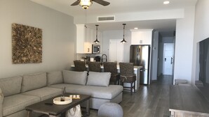 Een moderne woonkamer met een bank, salontafel en poefs. Achtergrond toont een keuken met een koelkast en magnetron.