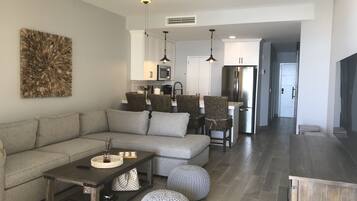 Un salon moderne avec un canapé, une table basse et des poufs. On aperçoit, au fond, un coin cuisine avec un réfrigérateur et un four à micro-ondes.