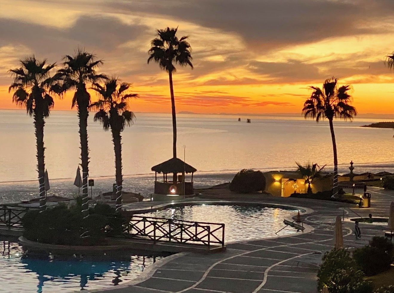 A resort pool area with palm trees, a gazebo, and a view of the sea at sunset.