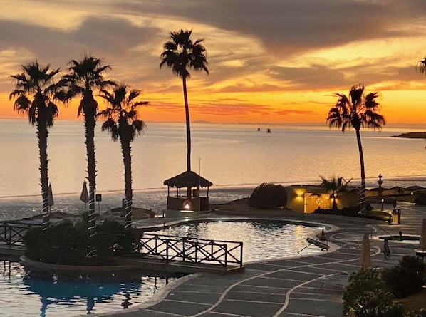 A resort pool area with palm trees, a gazebo, and a view of the sea at sunset.
