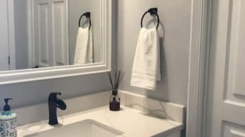 Ein Badezimmer mit einem weißen Waschbecken, einem schwarzen Wasserhahn und einem Spiegel. Zwei Handtücher hängen an Haken, eine Seifenspenderflasche und ein Schilferdiffusor sind vorhanden.