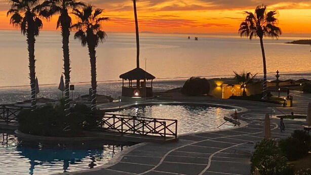 Zwembad van een resort met palmbomen, een prieel en uitzicht op de oceaan bij zonsondergang.