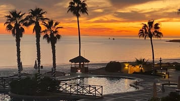 Un espace piscine d’un complexe hôtelier agrémenté de palmiers, d’un kiosque et offrant une vue sur l’océan au coucher du soleil.