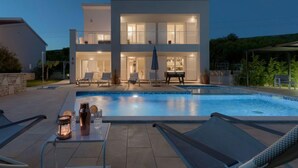 Ett modernt hus i två våningar med en swimmingpool, utomhusmöbler och en väl upplyst balkong.