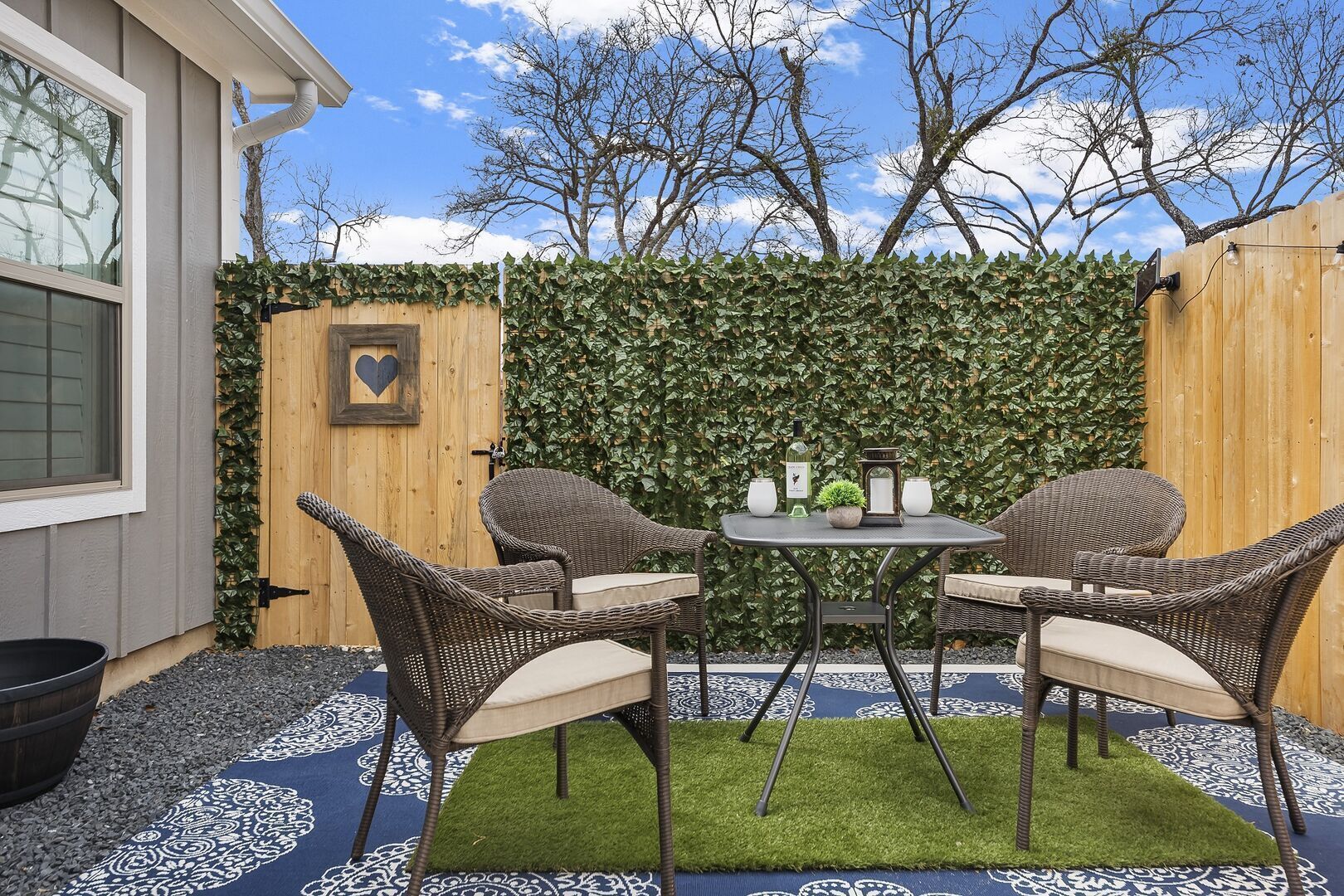 Een terras buiten met rieten meubels, een tafel en een hartvormige decoratie aan een houten hek.
