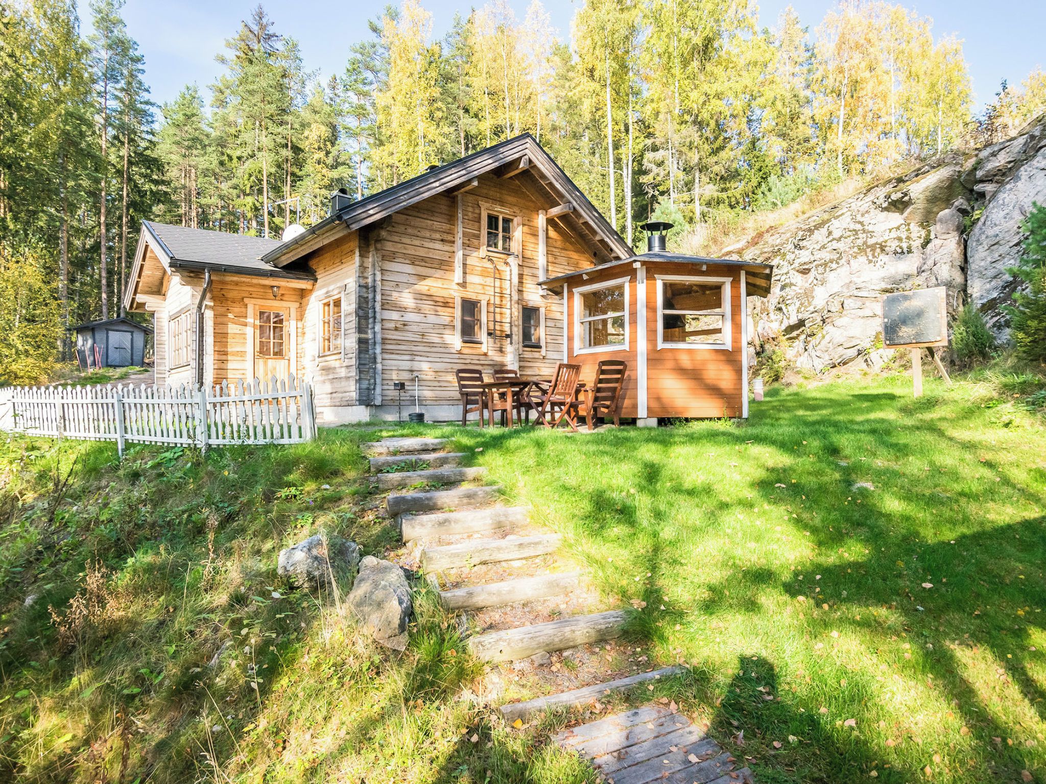 Una baita di legno con un portico e un'area pranzo, circondata da un recinto di legno bianco e un muro di pietra.
