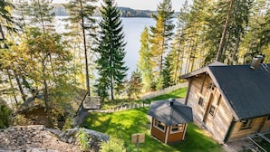 Une cabane avec une petite structure octogonale, entourée d’une clôture en bois, située près d’un lac bordé de grands arbres.