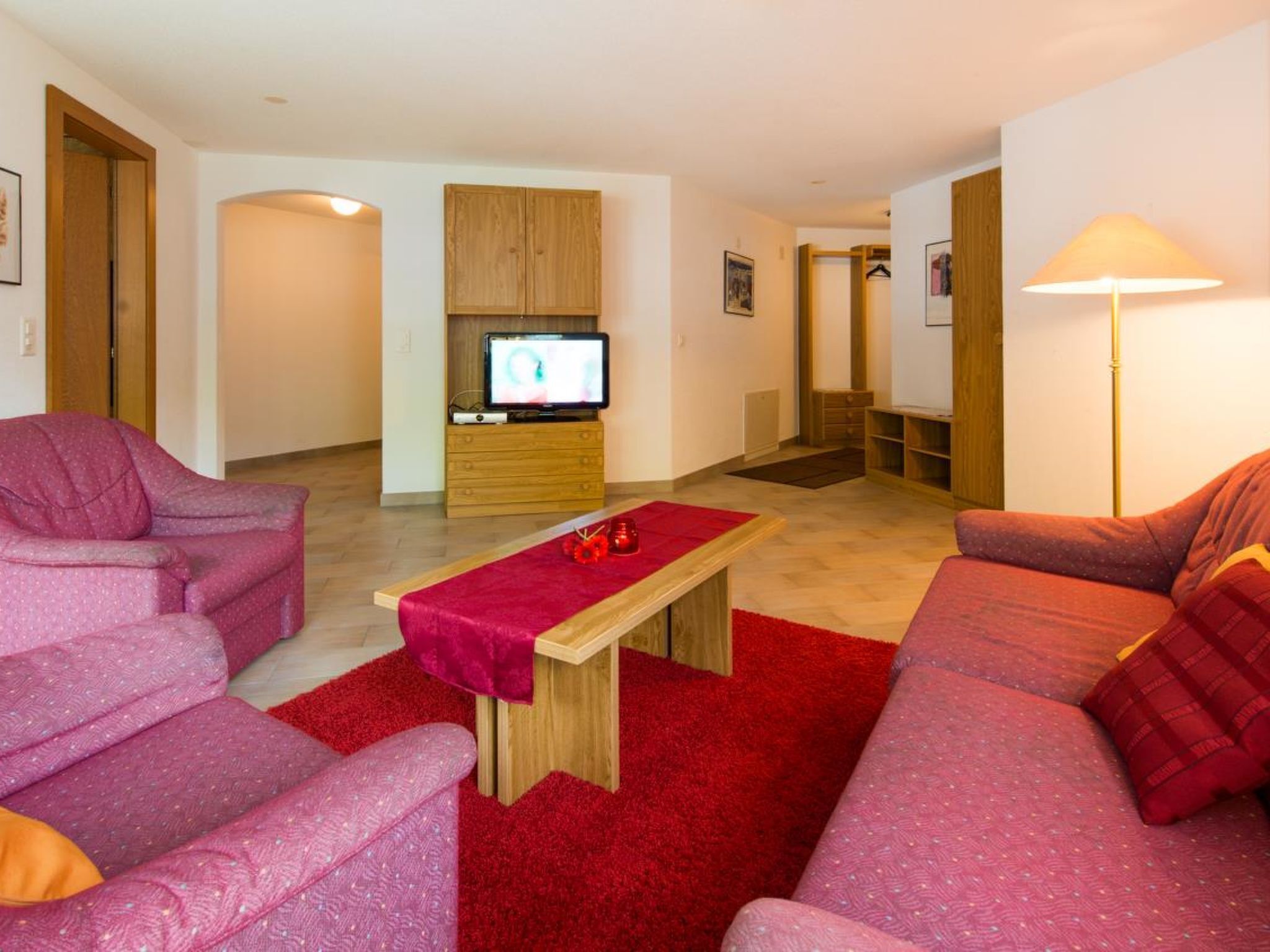 Un salon avec une moquette rouge, deux canapés roses, une table basse en bois, un téléviseur posé sur un meuble en bois et un lampadaire.