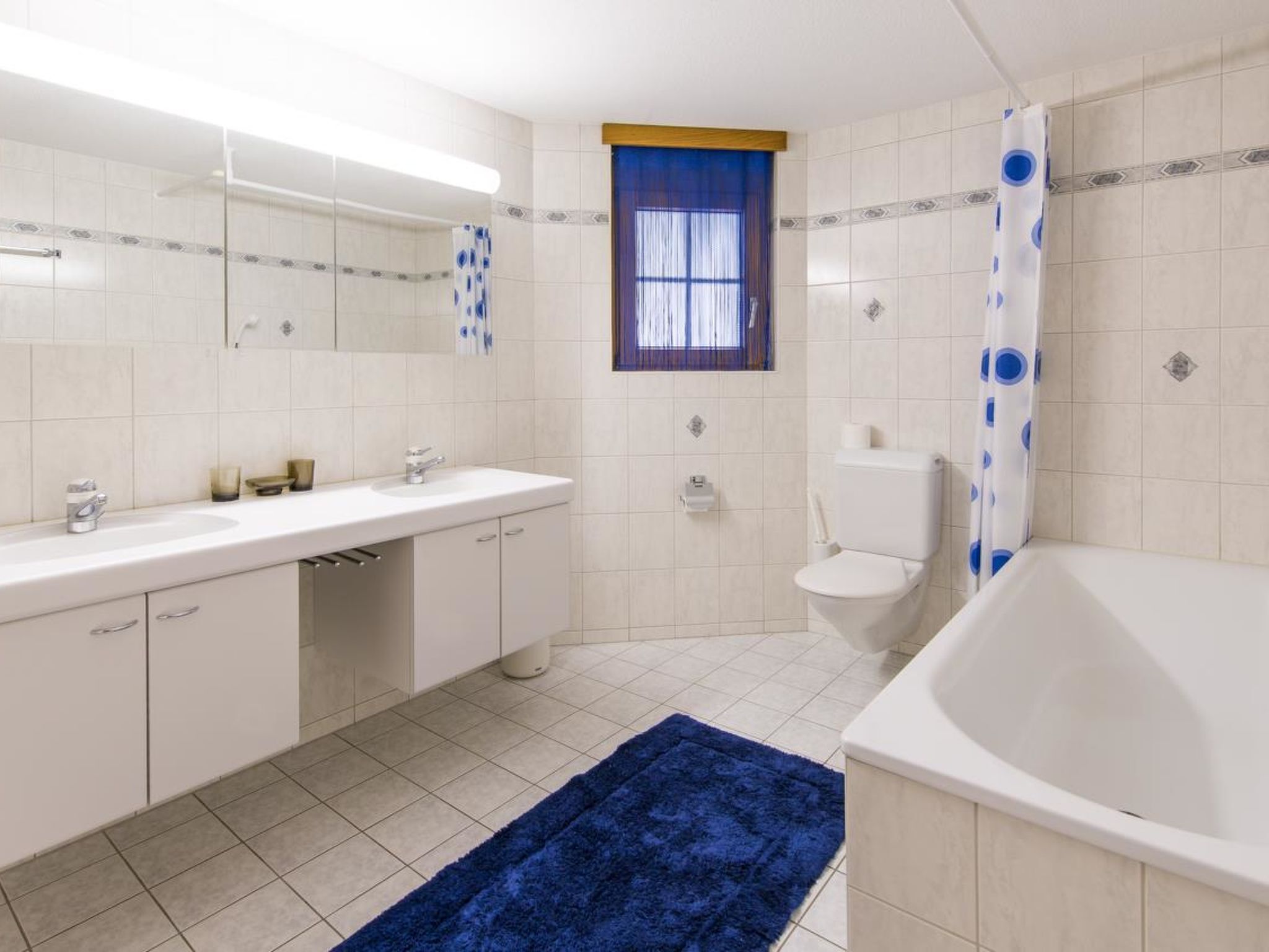 Un baño moderno con lavamanos, espejo y váter blancos. Hay una alfombra azul en el suelo y una cortina de ducha con diseño azul y blanco.