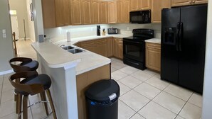 A modern kitchen with wooden cabinets, a black refrigerator, a microwave, and a sink. There are three bar stools and a trash can. - Walk to Super Bowl LV! Grand Spanish Style Home (Tampa)