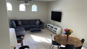 A living room with a gray sofa, a wooden dining table with chairs, a flat-screen TV on the wall, and a ceiling fan. - Walk to Super Bowl LV! Grand Spanish Style Home (Tampa)