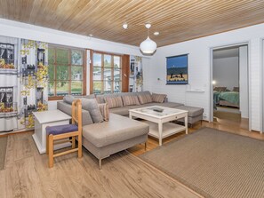 Ein modernes Wohnzimmer mit einer Eckcouch, einem Couchtisch und einem Esstisch. Es gibt ein großes Fenster mit Jalousien, eine Decke mit Holzlamellen und ein Gemälde an der Wand.