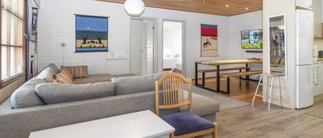 A modern living room with a grey sectional sofa, a wooden dining table, and a white refrigerator.