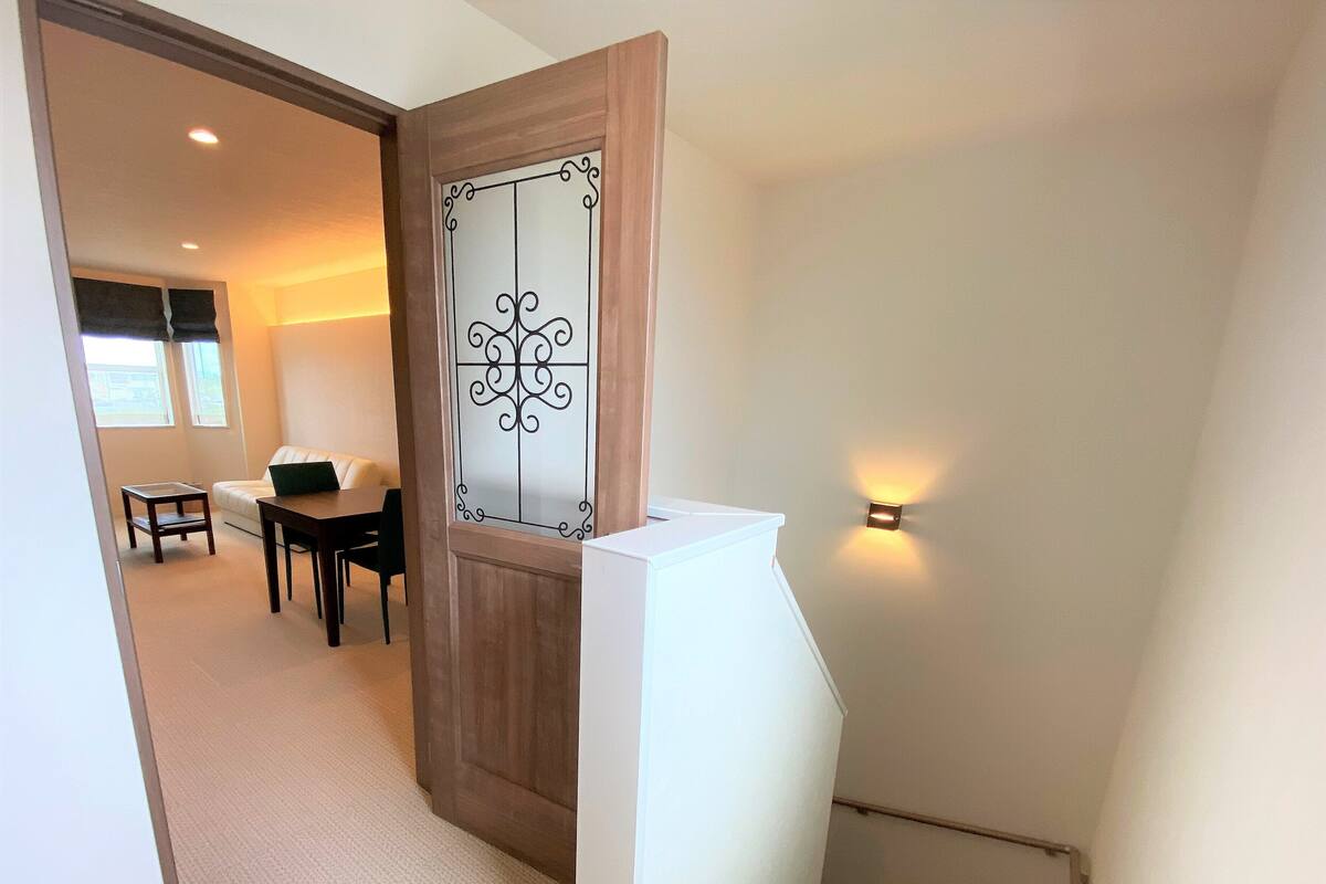 A hallway leading to a dining area with a white table and chairs, a wooden door with a decorative panel, and a wall-mounted light fixture.