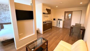 Un espacio habitable compacto con sofá, una mesa de centro de vidrio, televisor montado en la pared, kitchenette con microondas y refrigerador, y un área de comedor con una silla y mesa.
