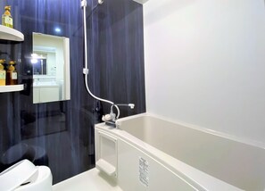 A modern bathroom with a bathtub, a shower, and a toilet. There is a mirror above the sink and a shelf with toiletries. - ecot Sunayama Beach (Miyakojima)