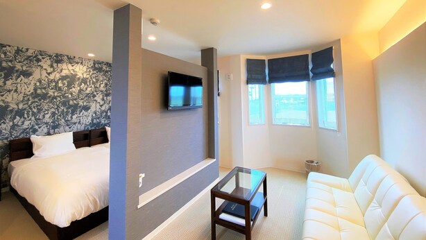 Una habitación de hotel con cama, sofá, televisor montado en la pared y ventana con cortinas.