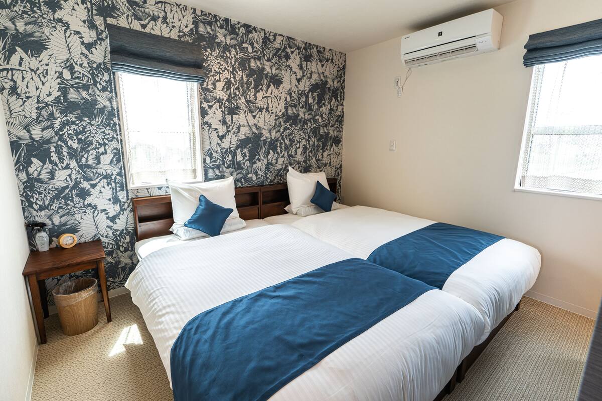 A double bed with blue and white bedding, a wooden nightstand with a lamp, a waste basket, and a wall-mounted air conditioner.