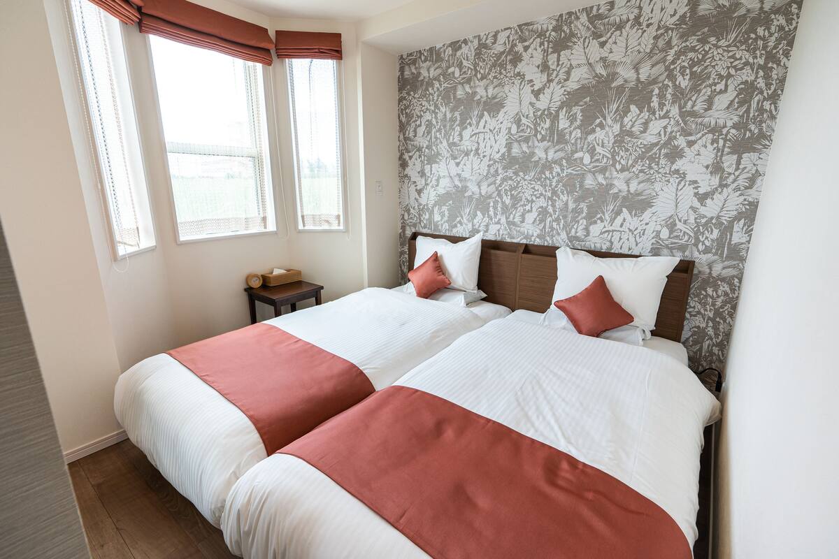 A double bed with white and red bedding, a wooden headboard, and a floral-patterned wall.