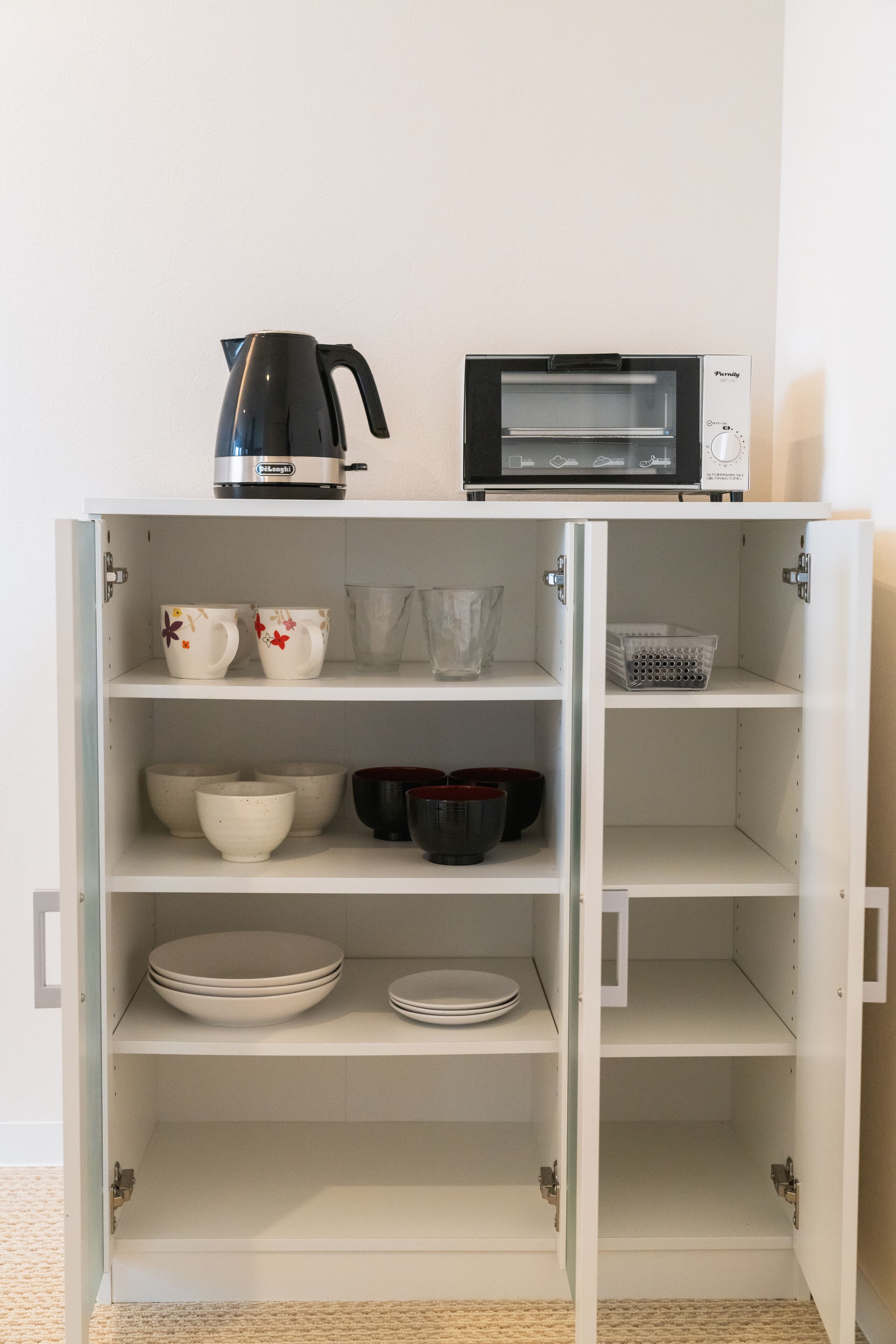 Un armario blanco con estantes abiertos que contiene utensilios de cocina, una tetera y un horno tostador.
