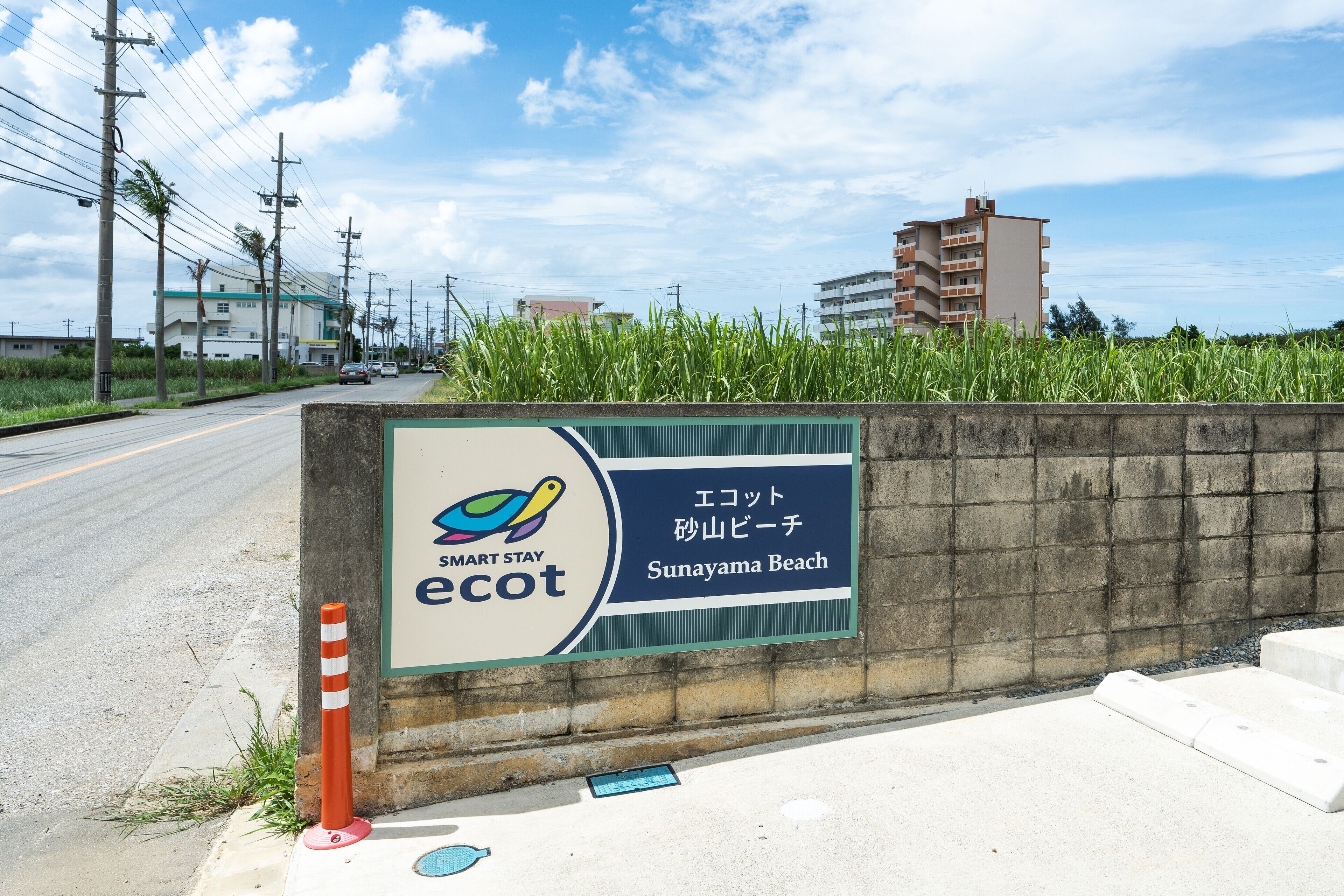 A sign for Smart Stay ecot Sunayama Beach with a concrete wall, a road, and buildings in the background.