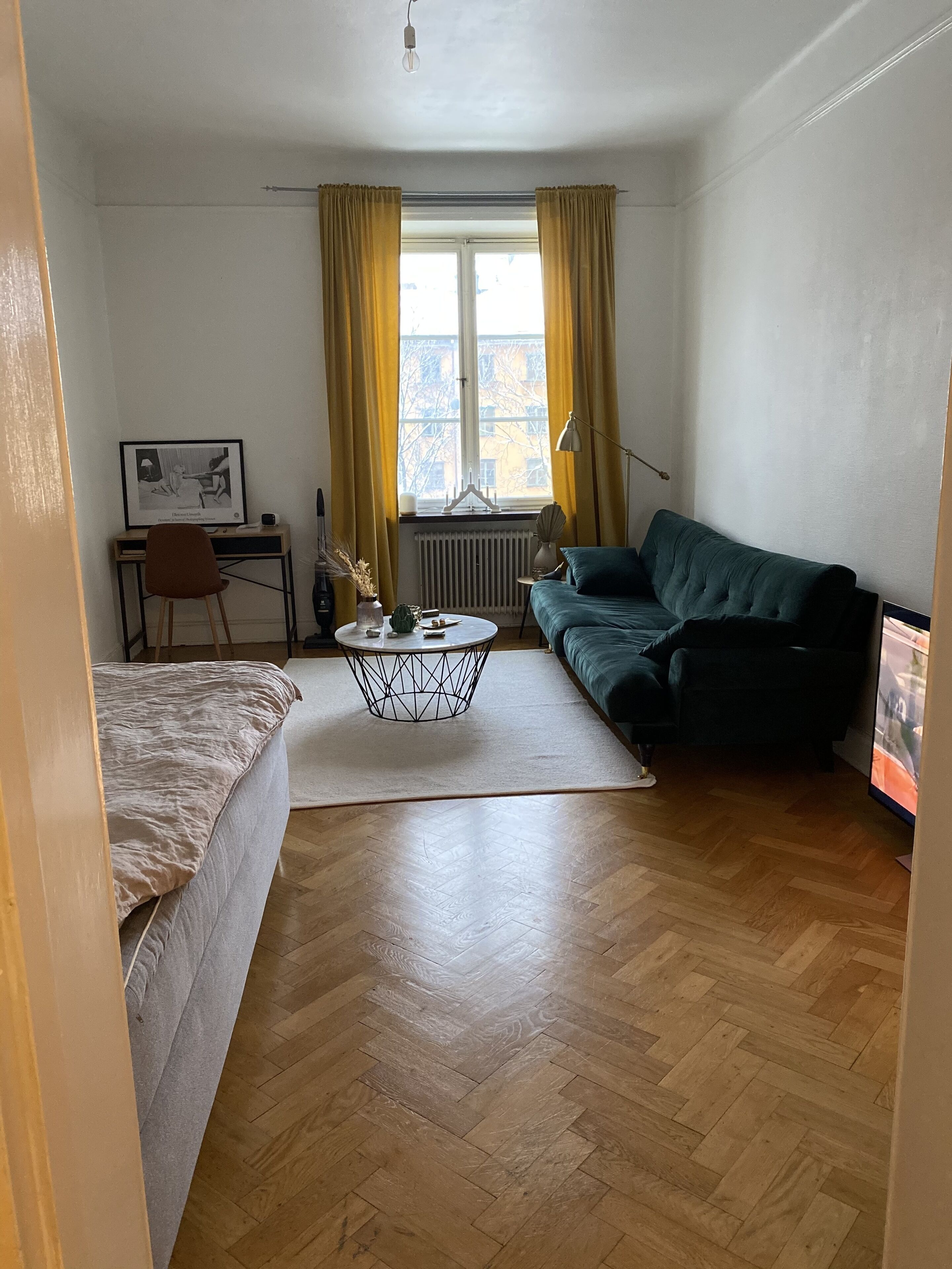Un espace de vie compact comprenant un lit, un canapé vert, une petite table basse ronde, un bureau avec une chaise et une fenêtre avec des rideaux.