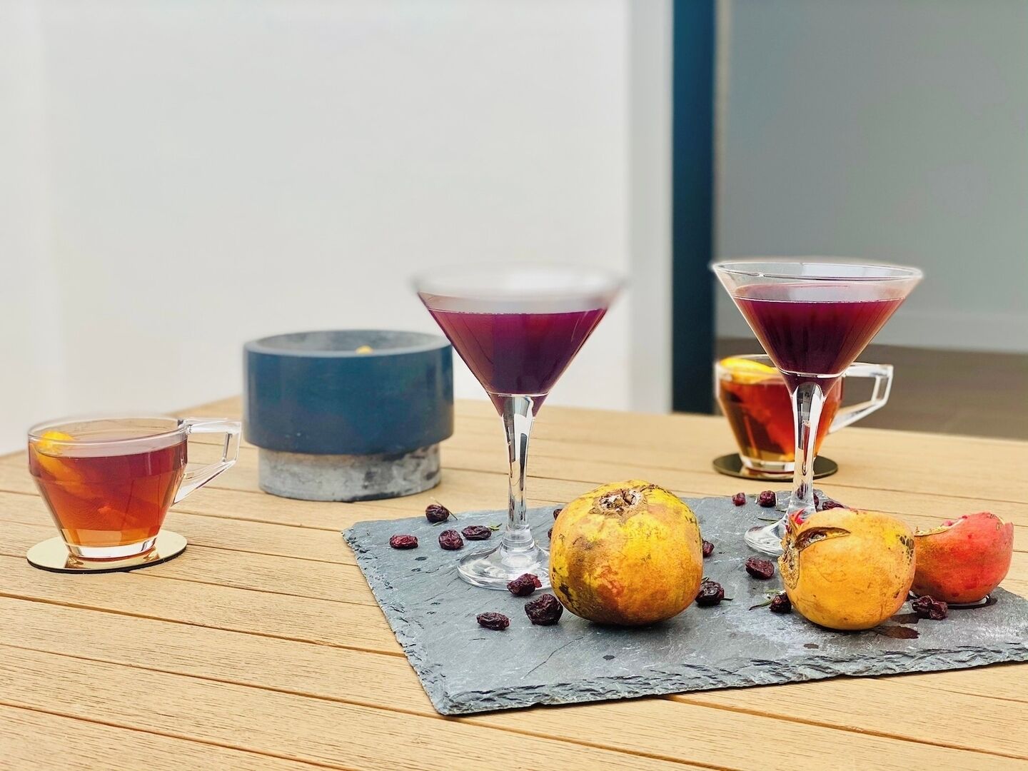 Una mesa con una taza de té, dos copas de martini con bebidas moradas, y dos frutas enteras y una partida sobre un posavasos de pizarra.