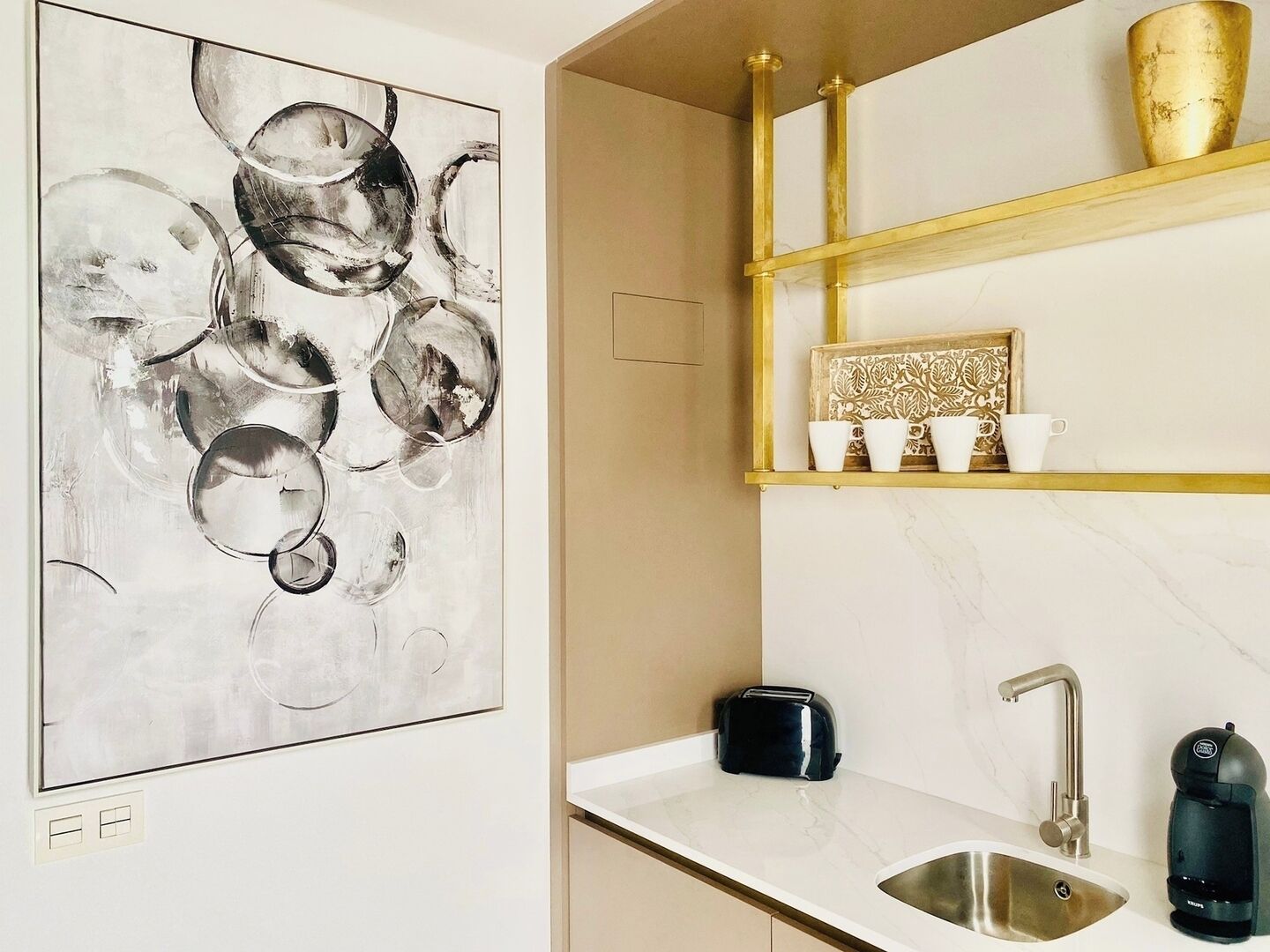 Una cocina moderna con encimera de mármol, un tostador negro, una cafetera y una estantería con marco dorado y tazas.