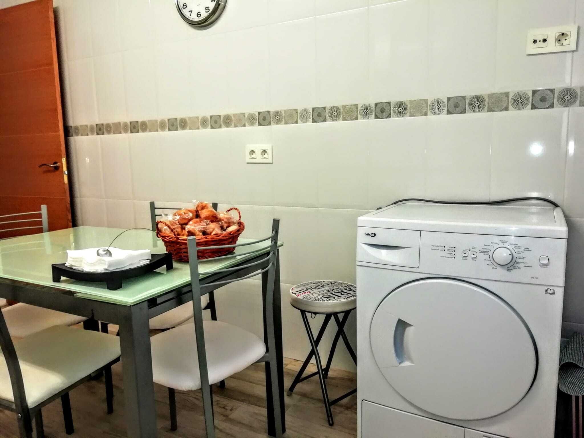 A small kitchen with a table, chairs, a basket of bread, a washing machine, and a clock on the wall.