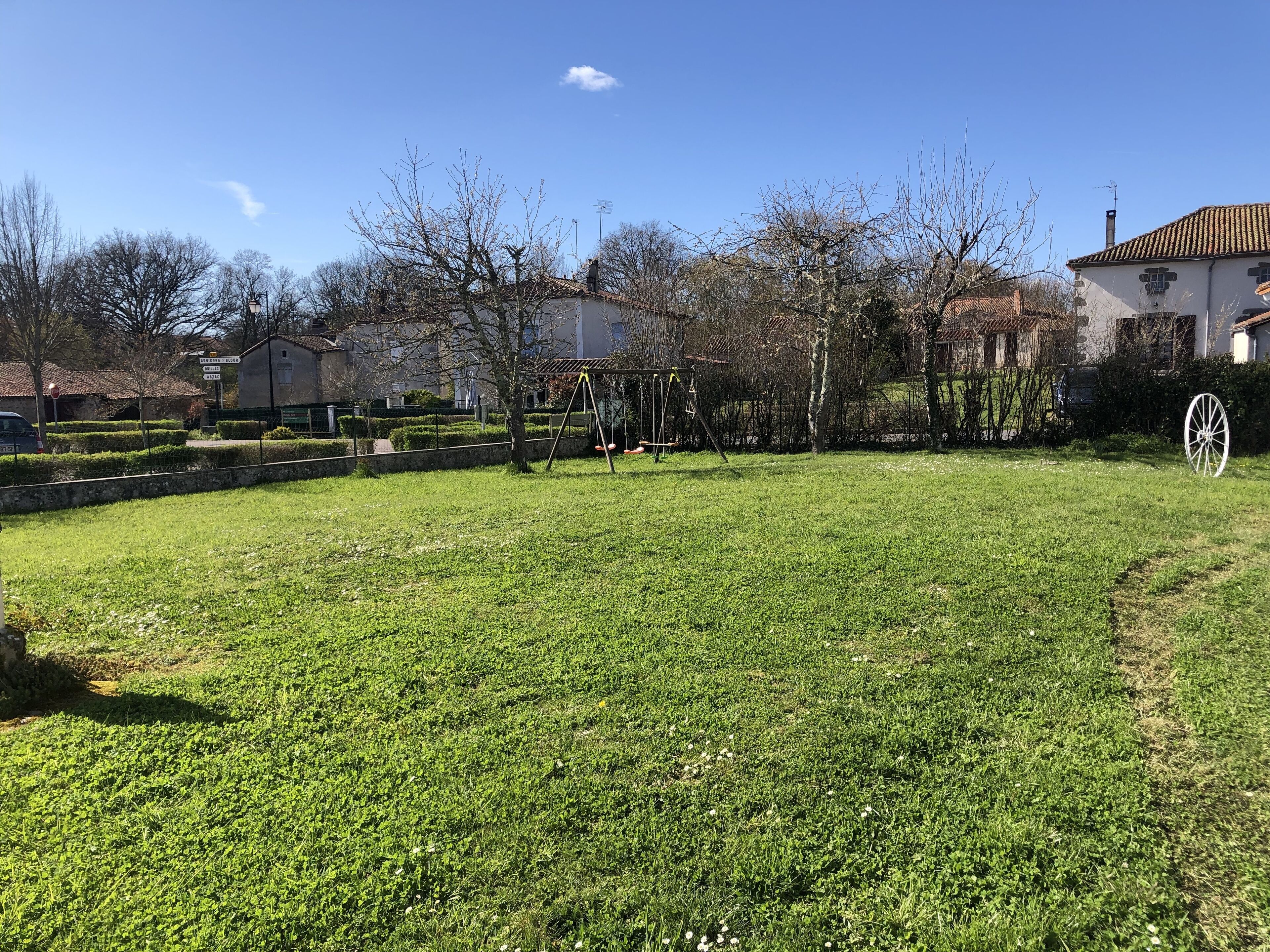 Ein grüner Garten mit Schaukel und einem weißen Gartenschubkarre, im Hintergrund ein Haus mit einem Ziegeldach.