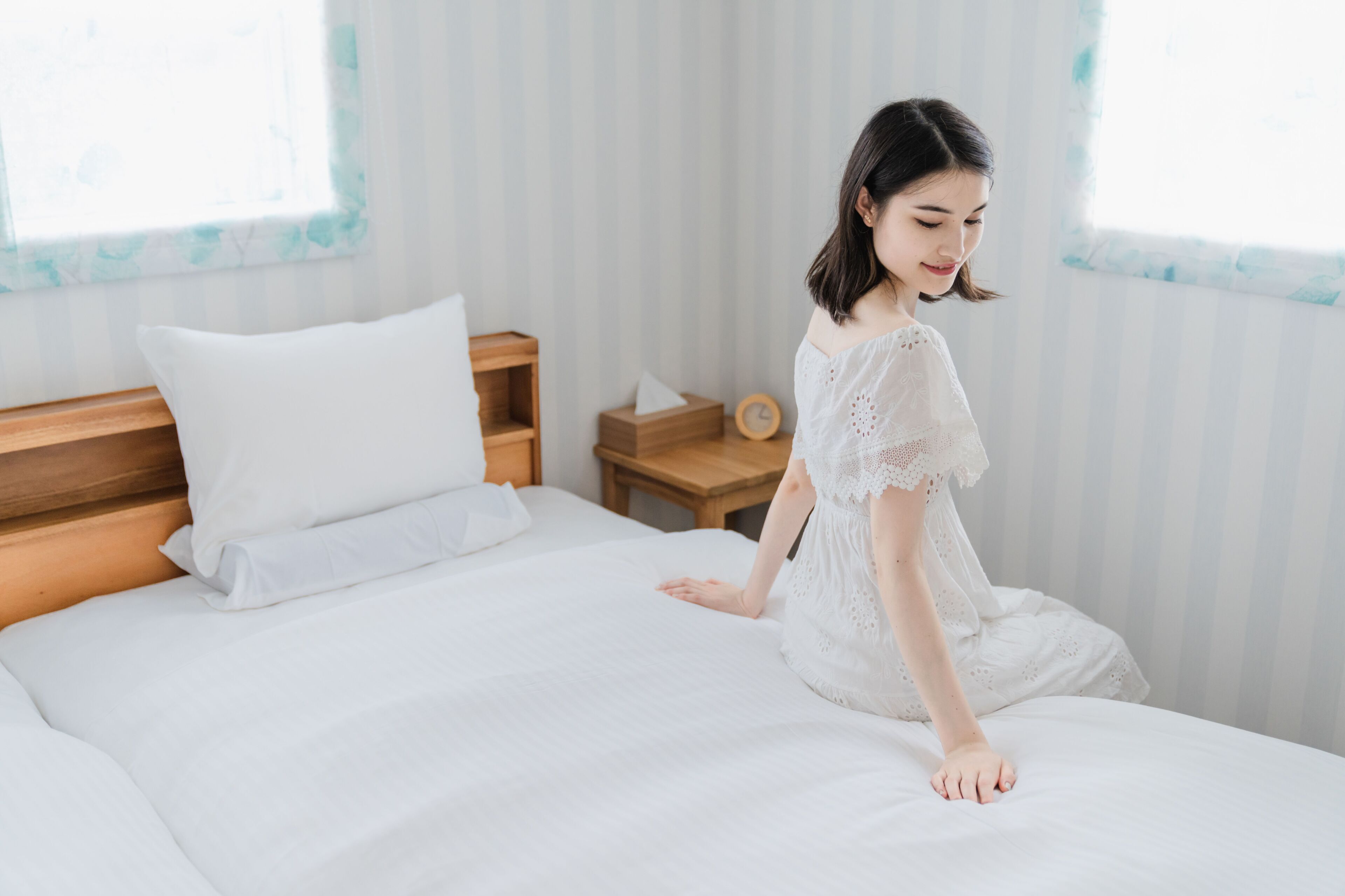 A bed with white linens, a wooden headboard, a bedside table with a clock, and a window with a view of trees.