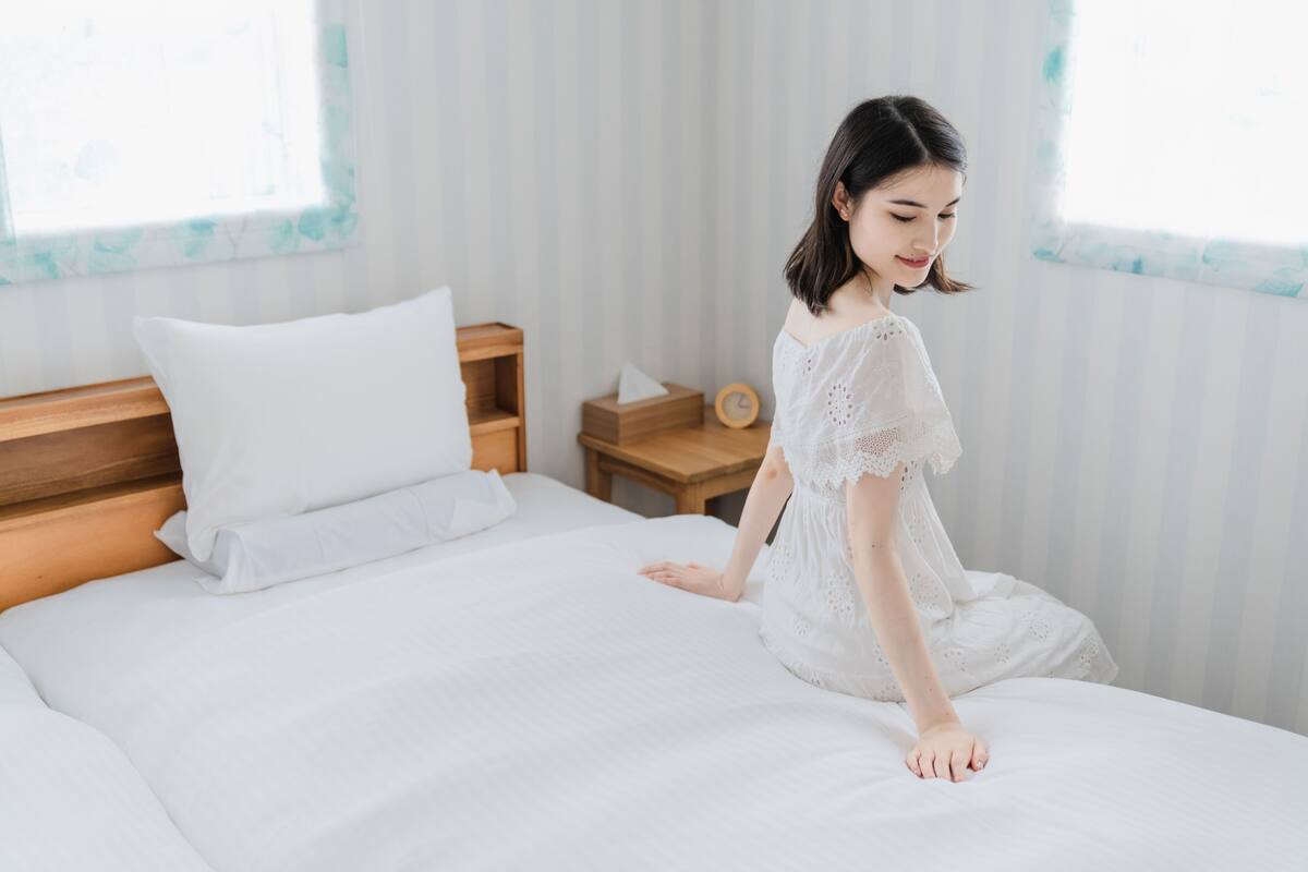A bed with white linens, a wooden headboard, a bedside table with a clock, and a window with a view of trees.